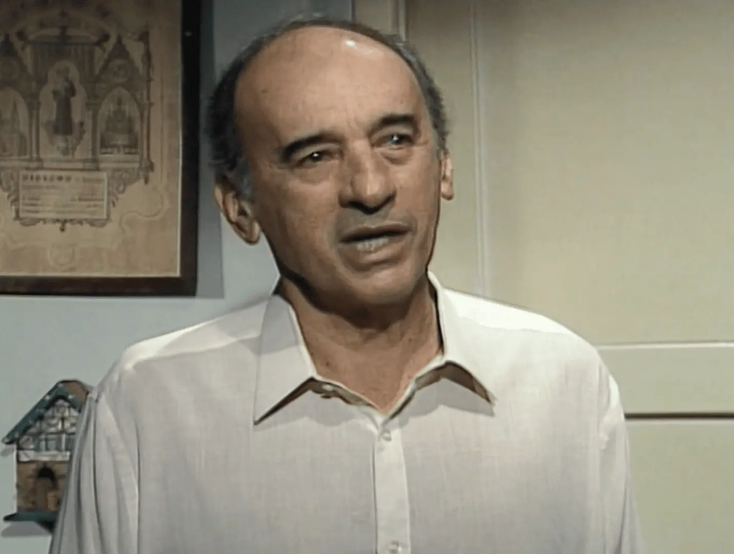 Personagem Salvador em Vale Tudo, homem mais velho, de pele clara, cabelos ralos e escuros nas laterais, aparece em ambiente interno usando uma camisa social clara. Ele está em discussão com a personagem Maria de Fátima. Ao fundo, vê-se uma parede com um quadro de moldura escura e uma pequena prateleira com um objeto decorativo em forma de casinha.