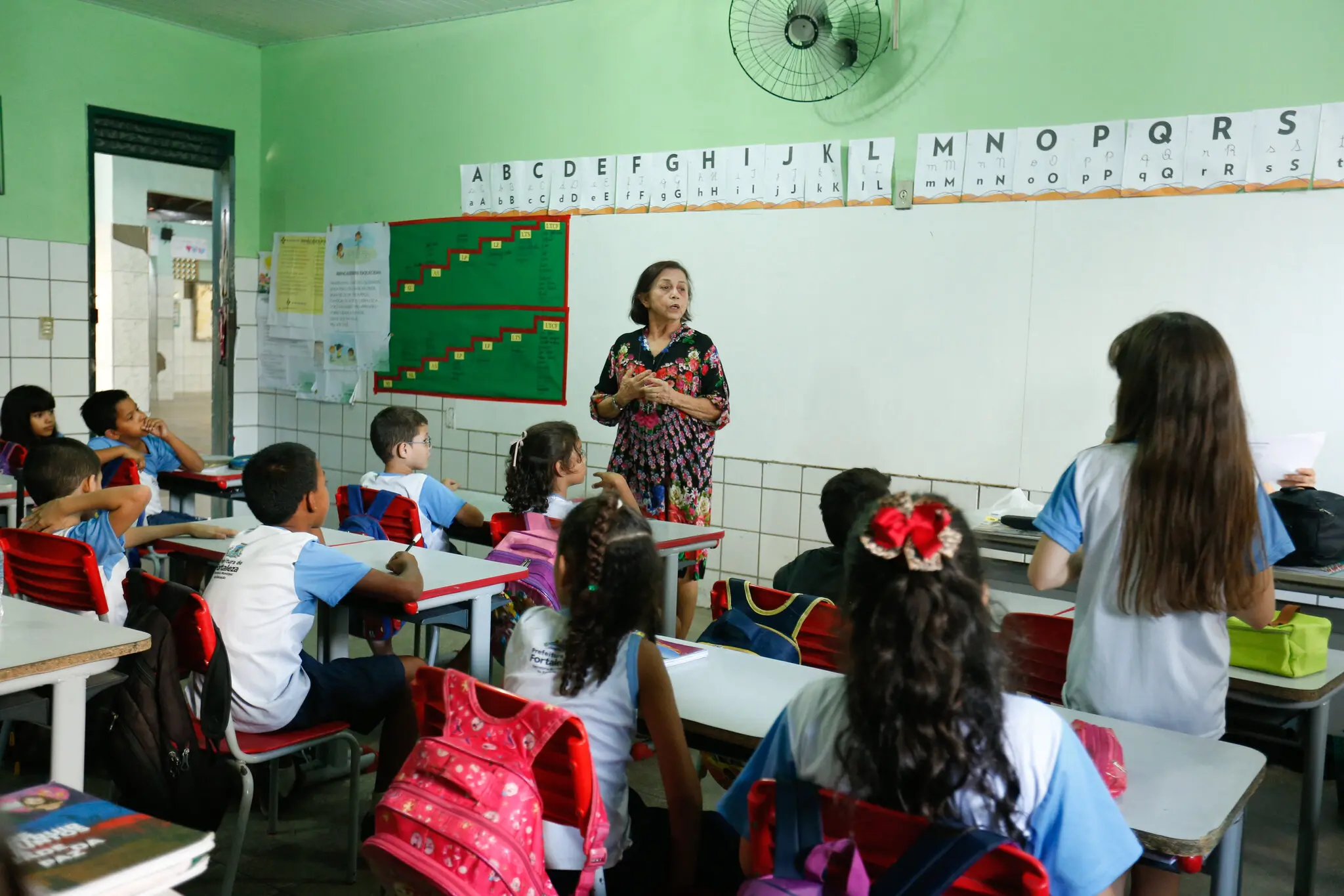 Sala de aula da rede municipal de ensino de Fortaleza. Crianças sentadas nas carteiras e uma mulher em pé, falando para a turma