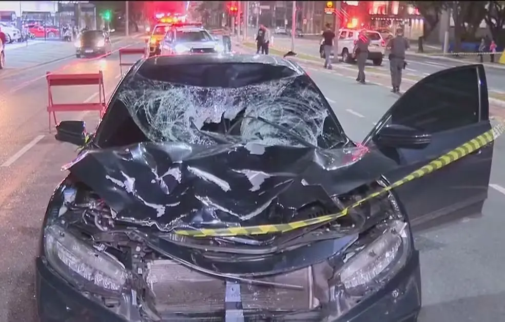 Imagem de carro acidentado com o capô amassado e vidro estilhaçado, evidenciando a gravidade do acidente. A cena mostra policiais e viaturas ao fundo, sinalizando um incidente sério.