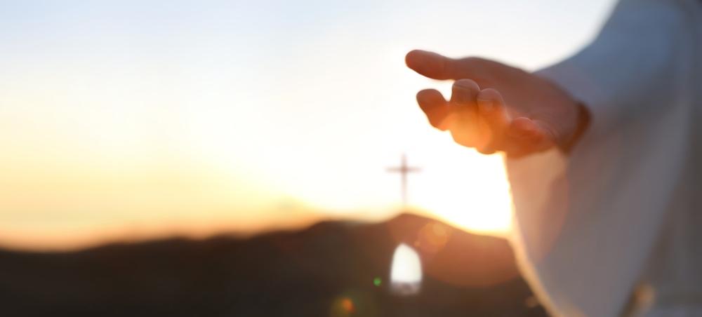 Foto de uma mão aberta em direção ao horizonte ao pôr do sol, simbolizando amor e acolhimento, com uma cruz visível ao fundo. Ideal para temas religiosos.