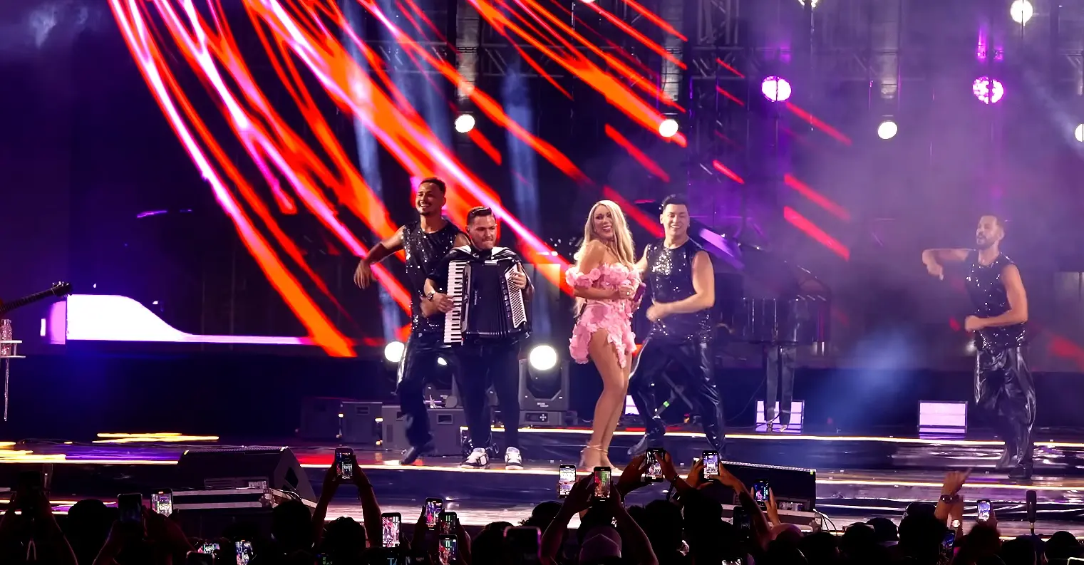 Cantora Taty Girl se apresenta no palco durante gravação de DVD em Fortaleza. Ela está ao centro, vestindo figurino rosa com babados e rodeada por quatro dançarinos com roupas pretas brilhantes. Ao lado esquerdo, um músico toca sanfona. O fundo do palco exibe luzes e efeitos visuais em vermelho e roxo. A plateia aparece em primeiro plano, com várias pessoas registrando o momento com seus celulares. O clima é de show vibrante e animado.