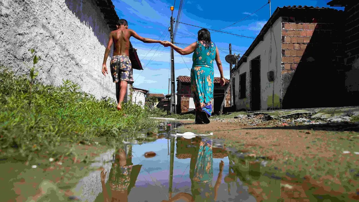 Uma mulher usa vestido longo, enquanto atravessa uma rua alagada em uma comunidade de Fortaleza. Ela segura a mão de um adolescente