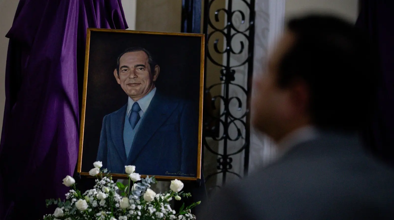 Igor Queiroz de perfil em primeiro plano desfocado e pintura de Edson Queiroz em segundo plano nítida, para matéria sobre missa de centenário de Edson Queiroz