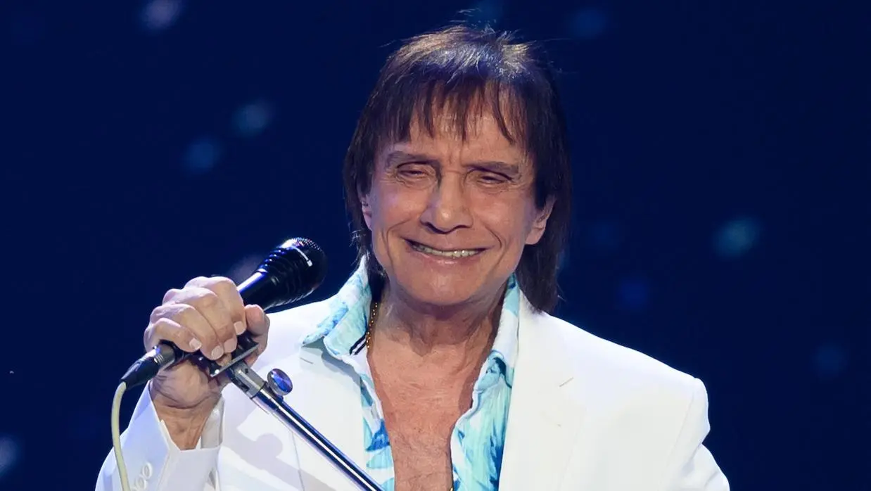 foto do cantor Roberto Carlos em show. Ele veste terno branco e blusa azul