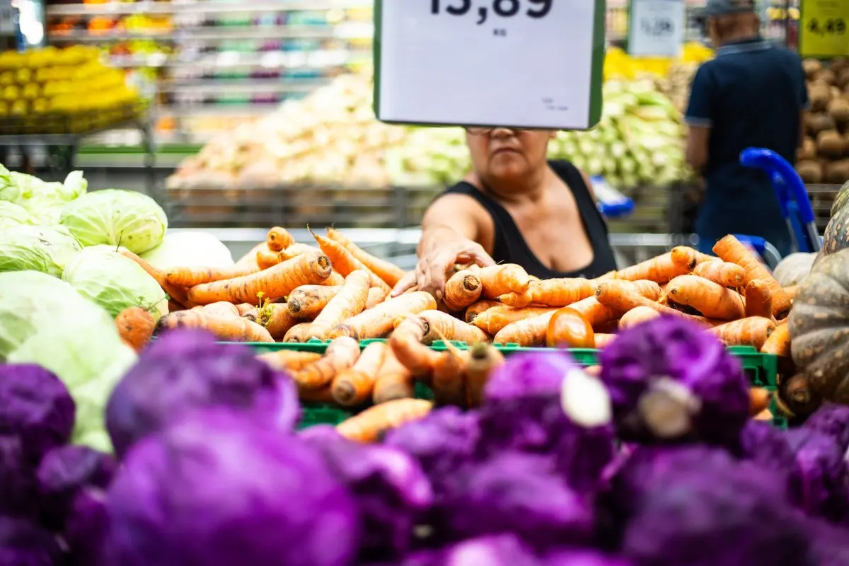 Close em verduras e legumes. Ao fundo, aparece uma consumidora pegando uma cenoura
