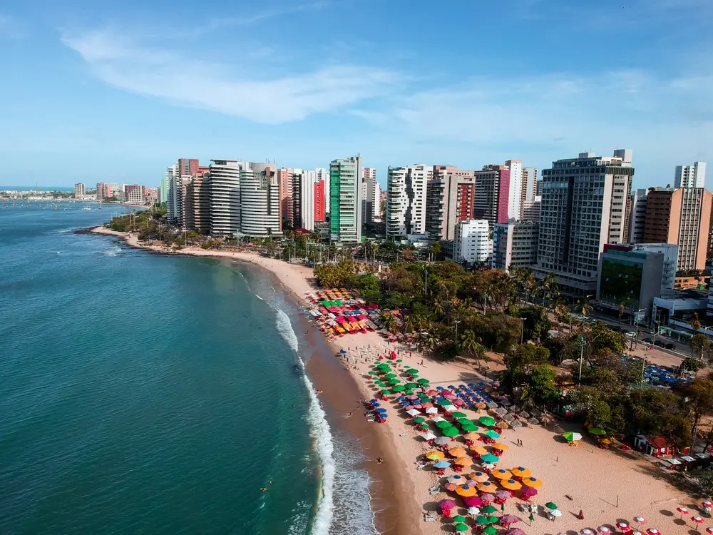 Foto de vista aérea da orla de Fortaleza, com prédios modernos ao fundo e diversas barracas de praia coloridas na areia.