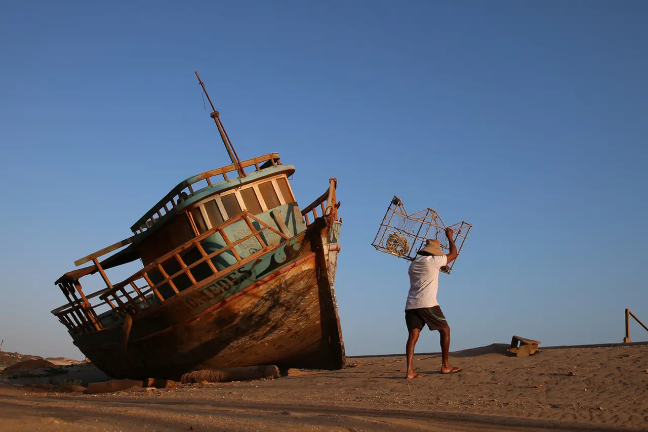 Imagem mostra um barco na areia e um pescador de lagosta carregando uma caçoeira, rede utilizada para pescar lagostas
