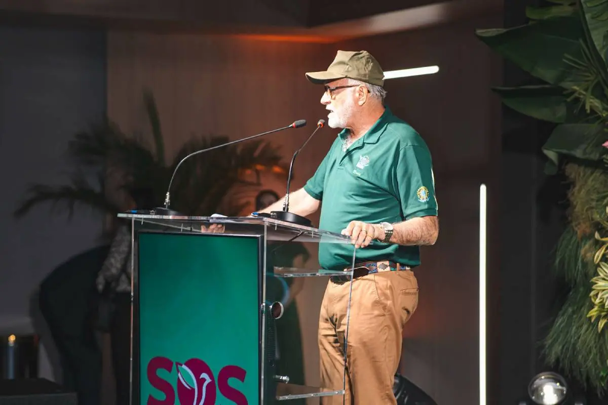 Deodato é um homem branco e grisalho, que veste fardamento do Ibama composto por blusa de gola polo verde e calça marrom, além de usar boné marrom e óculos de grau. Ele está ao microfone durante o evento, que tem decoração inspirada na serra, com folhas e muito verde