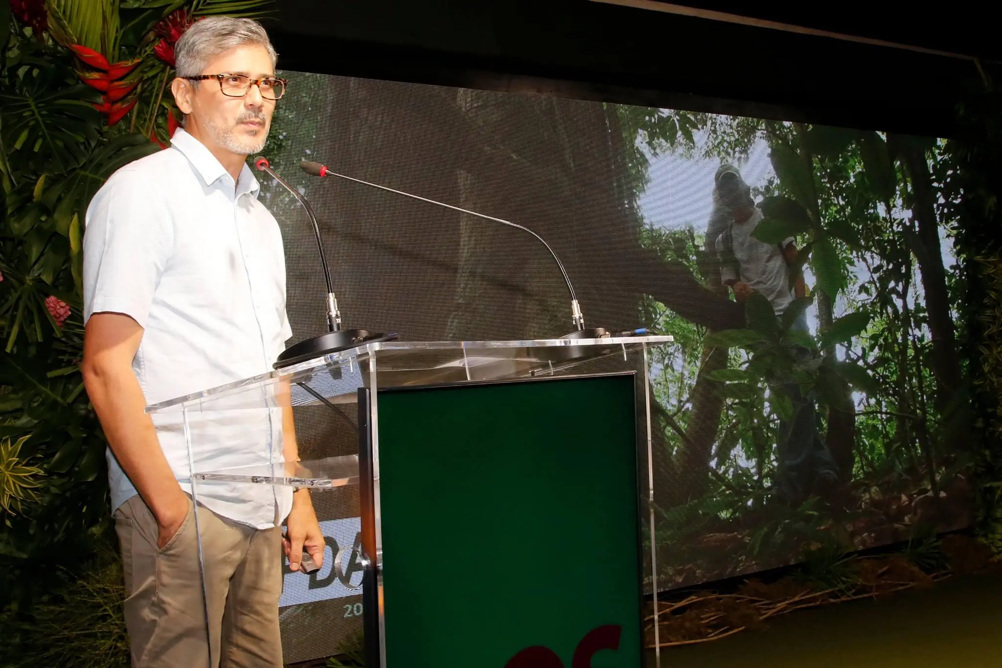 Um homem branco e grisalho, que veste camisa branca e calça bege e fala ao microfone durante o evento