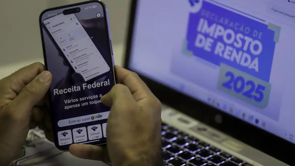 Imagem foca em mãos segurando celular aberto na página virtual da receita federal sobre tela de computador acessando o mesmo endereço para ilustrar lote de imposto de renda