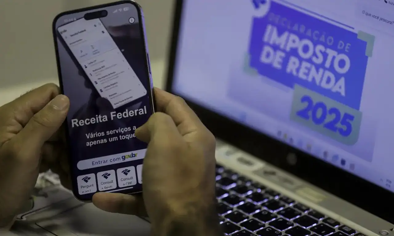 Imagem foca em mãos segurando celular aberto na página virtual da receita federal sobre tela de computador acessando o mesmo endereço para ilustrar lote de imposto de renda