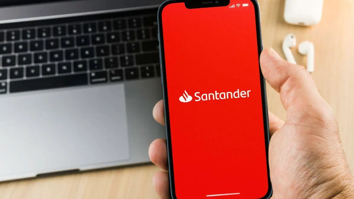 foto de celular com aplicativo do Santander na tela e um laptop ao fundo com fones de ouvido
