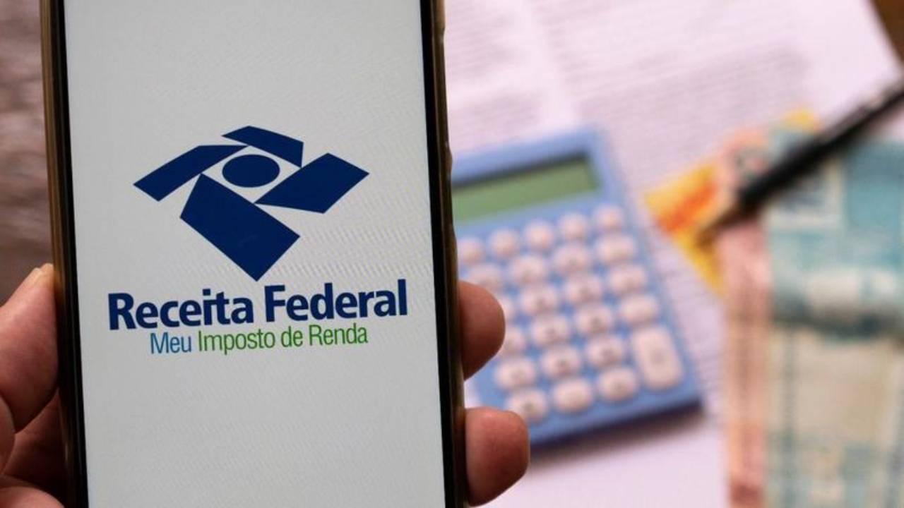 Celular mostrando aplicativo da Receita Federal em frente a mesa com documentos, caneta, calculadora e notas de Real ilustrando informações sobre isenção de imposto de renda por doença grave