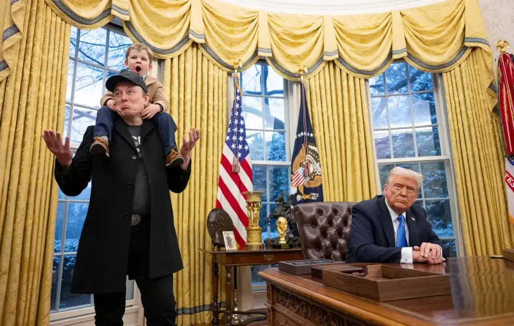 Elon Musk com o filho a tira colo, enquanto Trump fica sentado na mesa oficial do Salão Oval da Casa Branca