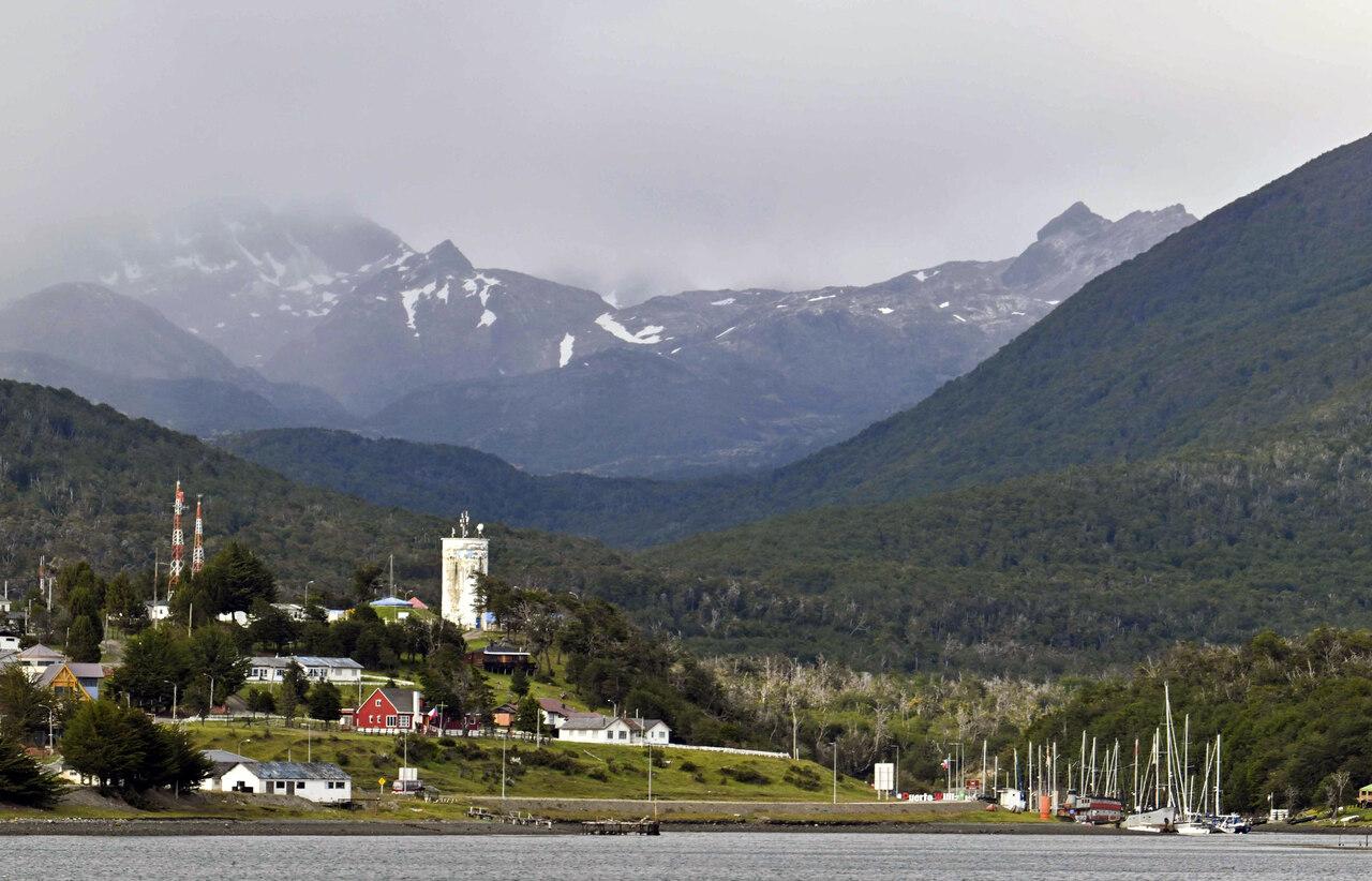 Vista da cidade chilena de Puerto Williams, cercada por montanhas e vegetação, com barcos atracados no porto e nuvens cobrindo parcialmente os picos ao fundo.