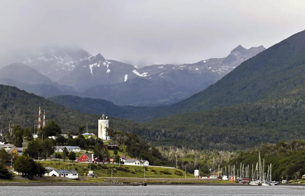 Vista da cidade chilena de Puerto Williams, cercada por montanhas e vegetação, com barcos atracados no porto e nuvens cobrindo parcialmente os picos ao fundo.