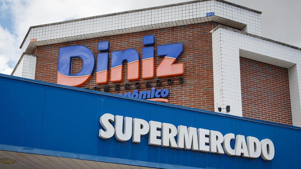 Foto que contém a fachada do Diniz Supermercados. Nome Diniz aparece personalizado em azul e laranja na parte de cima, enquanto o nome supermercado vem abaixo em branco em fundo azul.