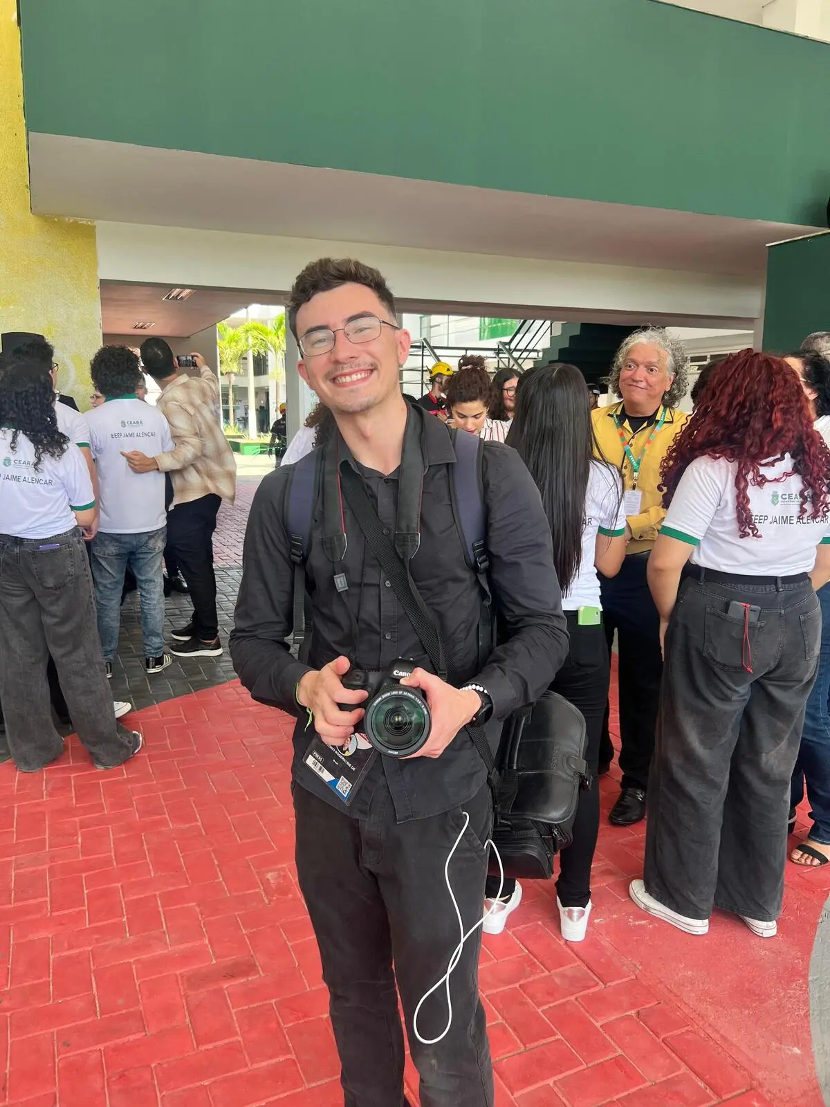 Rogério Bié veste preto e segura uma câmera fotográfica, enquanto sorri