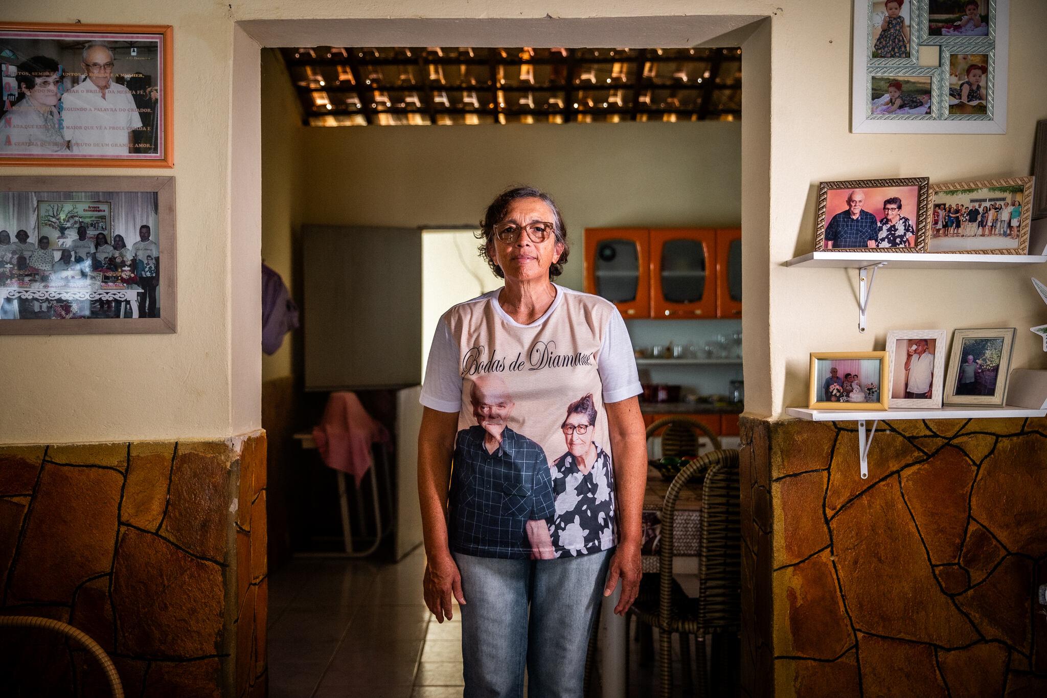 Mulher em pé, em casa, usando uma blusa com a foto dos pais. Ao redor dela, tem uma parede onde há porta-retratos com fotos de família