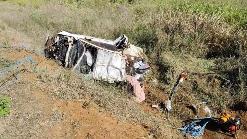 Imagem de um carro branco completamente destruído após capotar e cair em uma área de mato às margens de uma estrada de terra. O veículo está de lado, com partes da lataria e motor expostas. Há objetos e ferramentas espalhados ao redor. A vegetação ao redor é alta, indicando zona rural ou estrada pouco movimentada. Cena típica de um grave acidente de trânsito