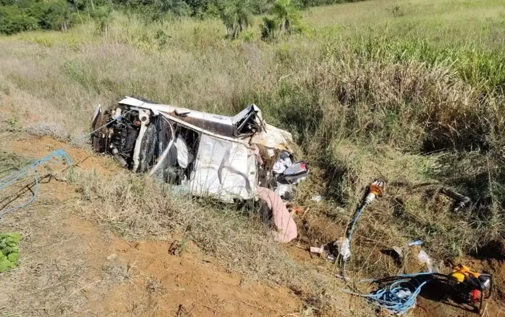 Imagem de um carro branco completamente destruído após capotar e cair em uma área de mato às margens de uma estrada de terra. O veículo está de lado, com partes da lataria e motor expostas. Há objetos e ferramentas espalhados ao redor. A vegetação ao redor é alta, indicando zona rural ou estrada pouco movimentada. Cena típica de um grave acidente de trânsito