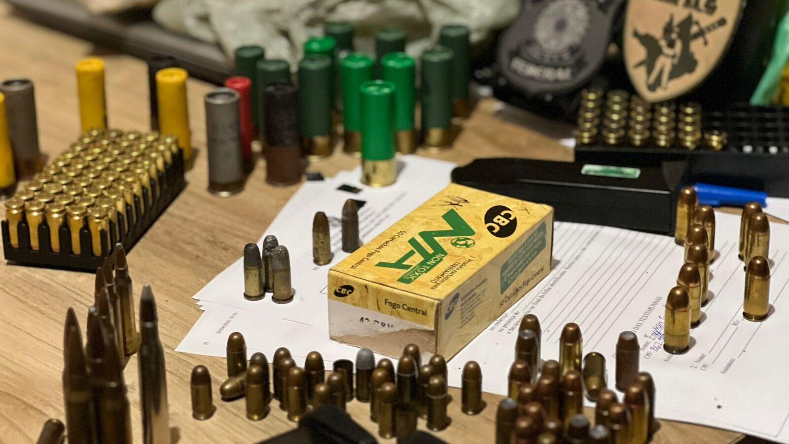 A imagem mostra uma mesa repleta de munições de diferentes calibres, incluindo cartuchos de espingarda, balas de pistola e munições de fuzil. No centro da imagem, há uma caixa amarela da marca CBC (Companhia Brasileira de Cartuchos), identificada como 