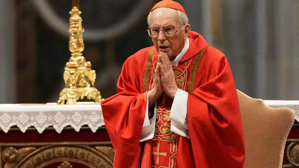 Cardeal em vestes religiosas, realizando um gesto de oração em uma cerimônia no Vaticano. Imagem capturada durante um evento importante da Igreja Católica.