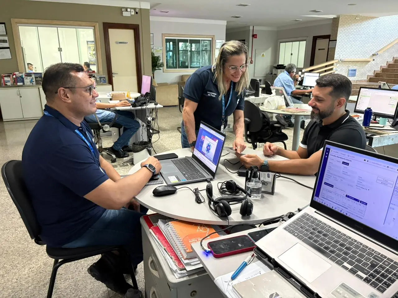 Foto no cenário de trabalho em que dois funcionários, sendo uma mulher e dois homens, aparecem sorrindo enquanto trabalham