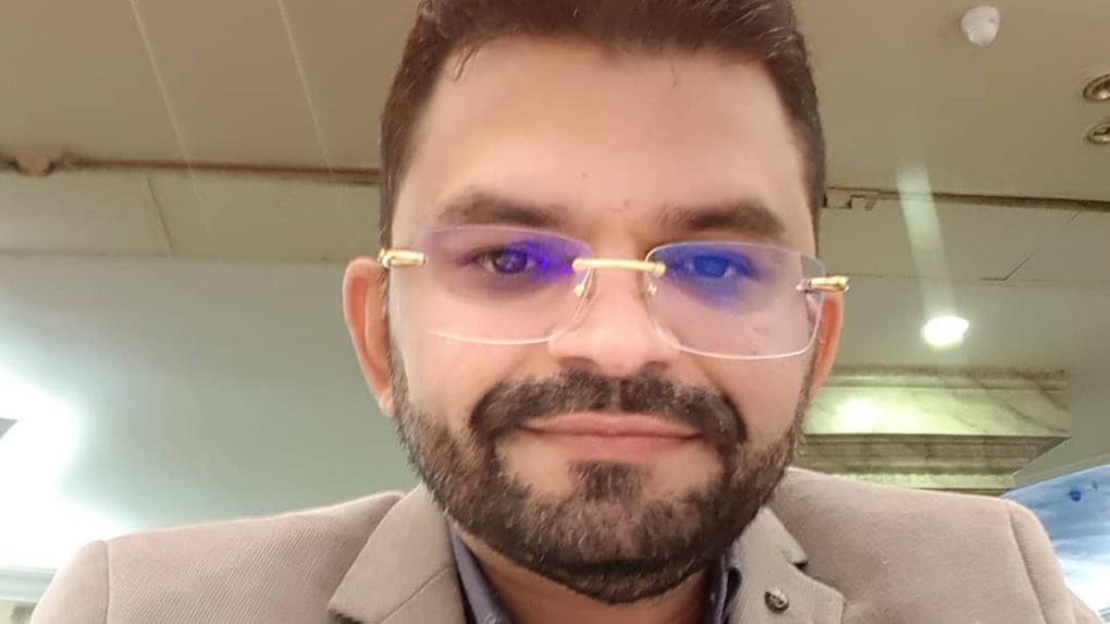 Vinícius Cunha Batista, dono de rádio morto em saída de padaria. Ele tem pele clara, barba cheia e cabelos curtos e escuros, usando óculos com lentes levemente azuladas e armação dourada. Ele veste um blazer bege claro sobre uma camisa azul. Está sorrindo levemente, em ambiente interno com iluminação artificial e teto com forro aparente.