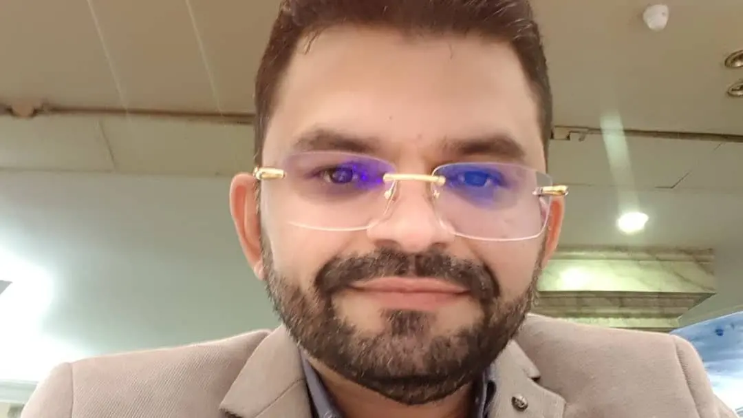 Vinícius Cunha Batista, dono de rádio morto em saída de padaria. Ele tem pele clara, barba cheia e cabelos curtos e escuros, usando óculos com lentes levemente azuladas e armação dourada. Ele veste um blazer bege claro sobre uma camisa azul. Está sorrindo levemente, em ambiente interno com iluminação artificial e teto com forro aparente.