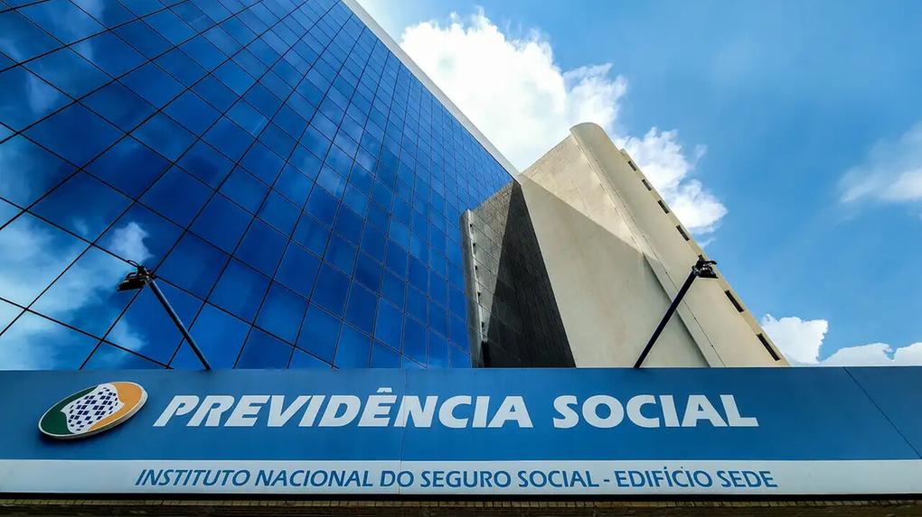 A imagem mostra o edifício sede do Instituto Nacional do Seguro Social (INSS), com sua fachada de vidro azul refletindo o céu. O prédio possui uma estrutura moderna e imponente, com uma placa na entrada indicando o nome da instituição.