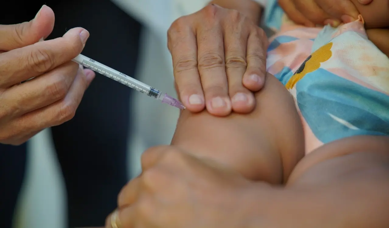 Profissional de saúde aplica injeção em bebê; a seringa é posicionada na coxa da criança, enquanto mãos seguram a perna para estabilização durante a vacinação
