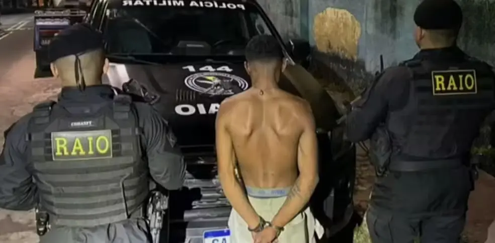 Suspeito aparece de costas e sem camisa enquanto é conduzido por dois policiais para uma viatura