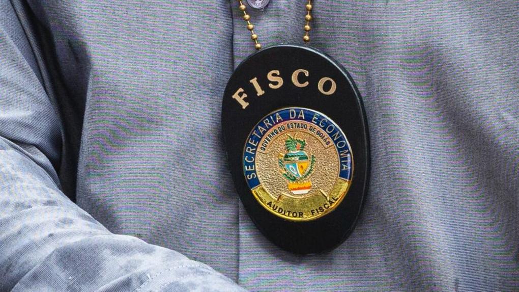 Imagem mostra o torso de um agente com camisa azul e distintivo de auditor fiscal. Sefaz GO lança concurso com 300 vagas
