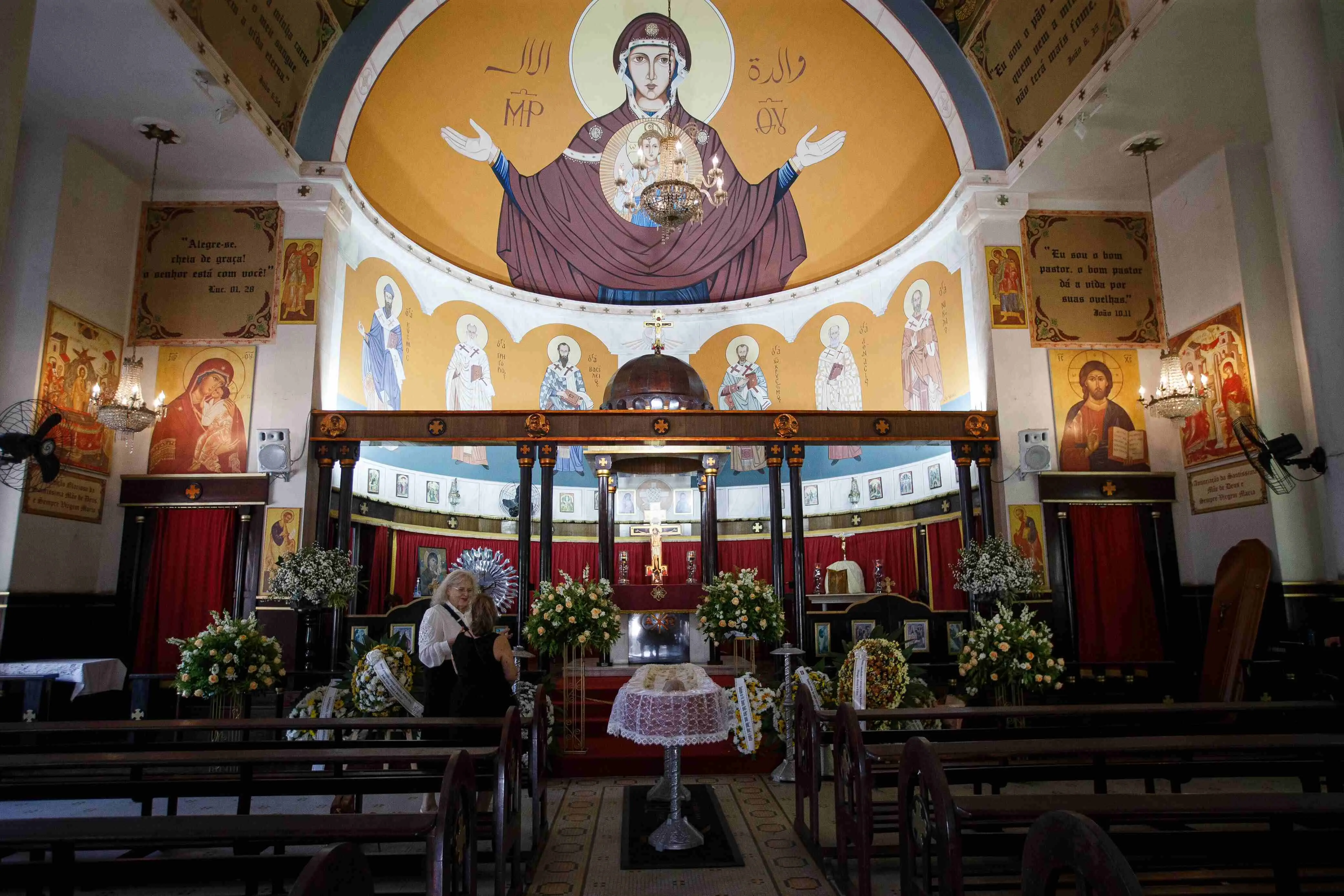Interior da Igreja do Líbano, em Fortaleza de rito maronita no Líbano, decorada com ícones religiosos de tradição oriental. No centro da imagem há um altar adornado com flores brancas e amarelas, uma cruz dourada e cortinas vermelhas. O teto semicircular apresenta uma grande imagem da Virgem Maria com os braços abertos, ladeada por inscrições em árabe e grego, além de outras figuras de santos. Em primeiro plano, dois bancos e uma mesa redonda coberta por toalha de renda. Duas mulheres vestidas de preto estão próximas ao altar, indicando um velório. Ao fundo, vê-se um caixão cercado por coroas de flores.