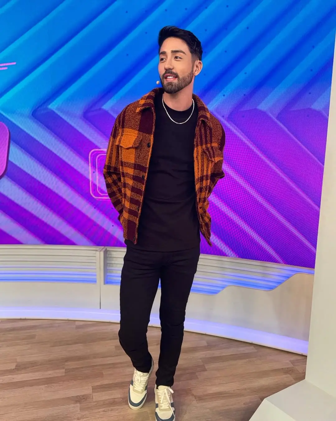 Influenciador Rico Melquíades com casaco xadrez laranja e preto posando em estúdio colorido, exibindo estilo moderno e despojado.