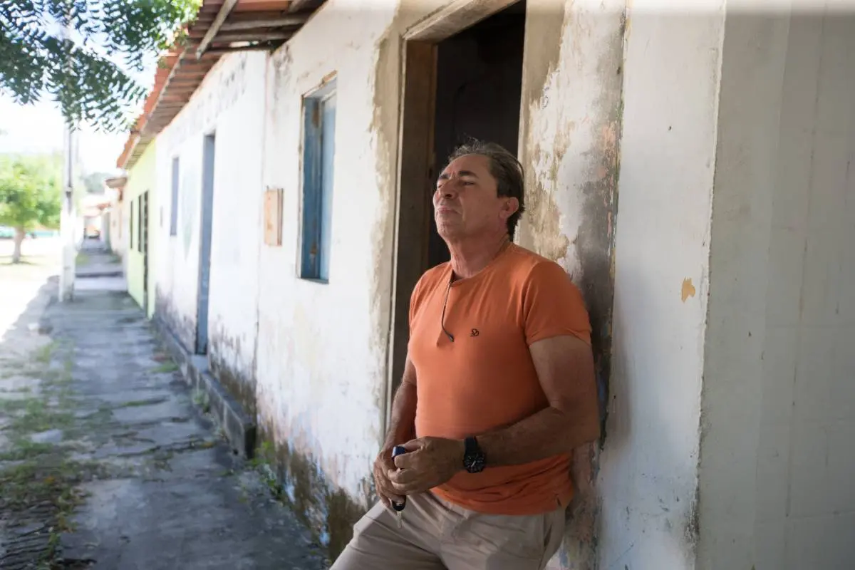 Morador encostado em uma casinha antiga. Ele é um home branco, grisalho e veste uma camisa laranja