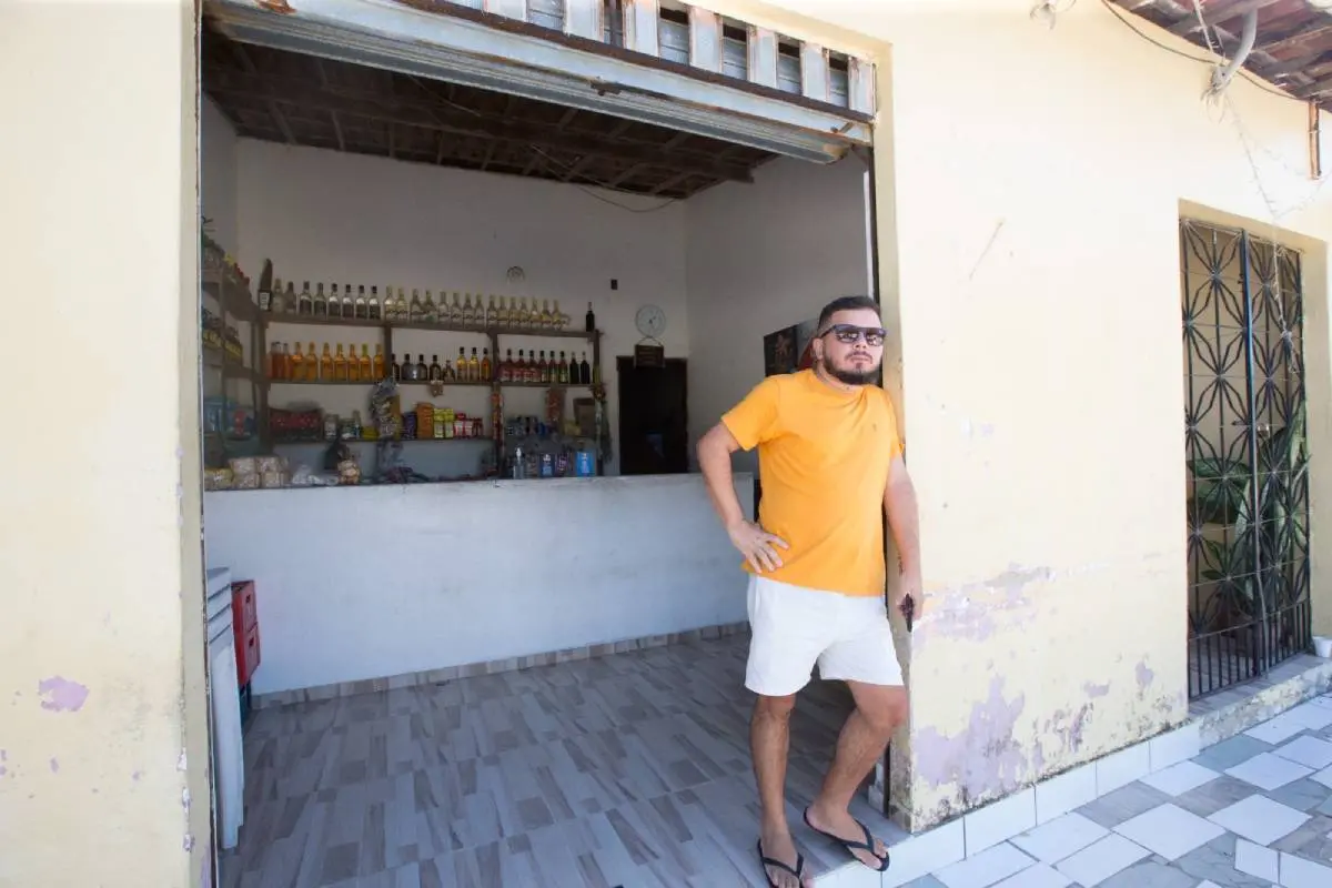 Um homem jovem encostado na muro da lojinha. Ele veste uma camisa amarela e short branco, além de usar óculos escursos