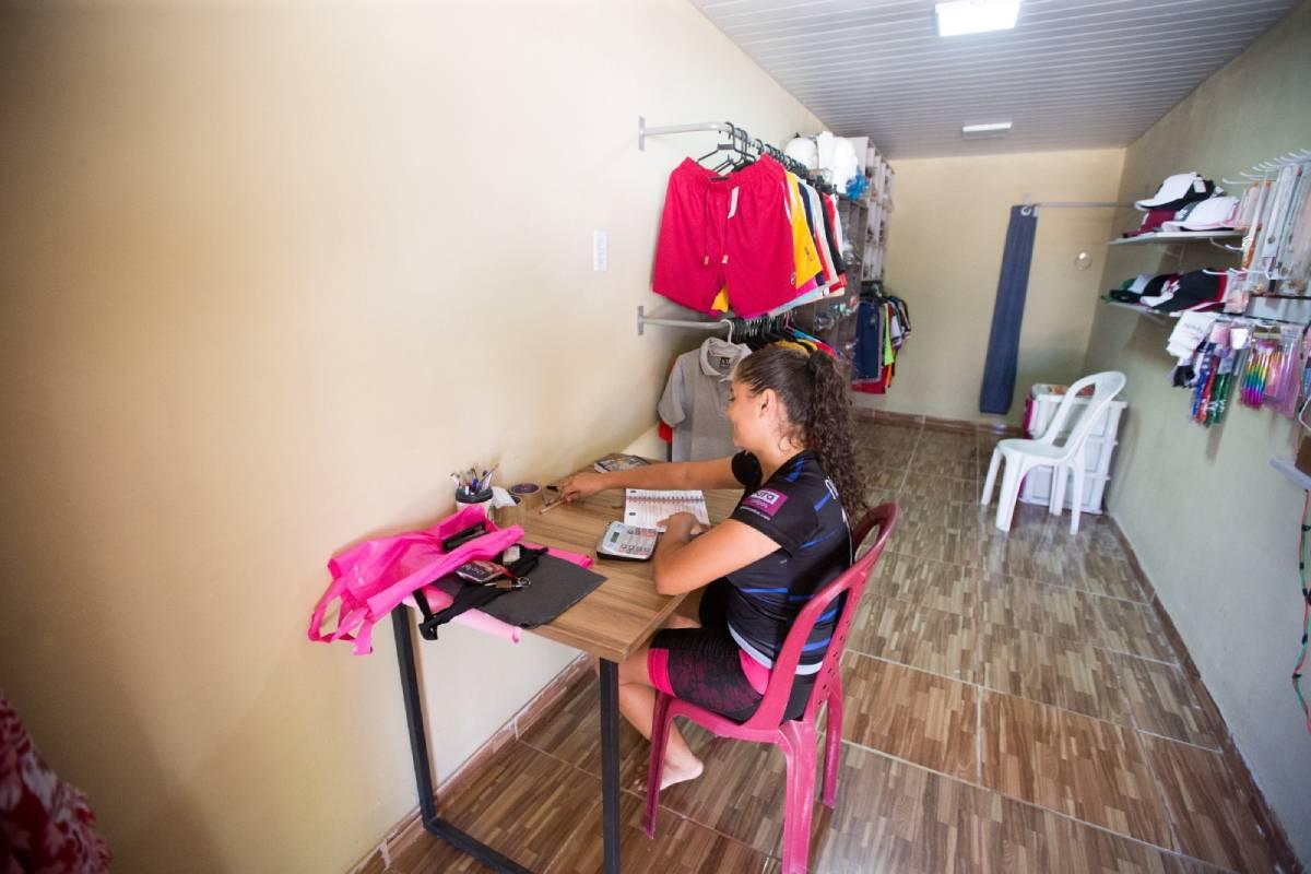 Uma mulher está sentada de costas em uma escrivaninha. Ela tem cabelos cacheados e veste uma camisa de time de futebol