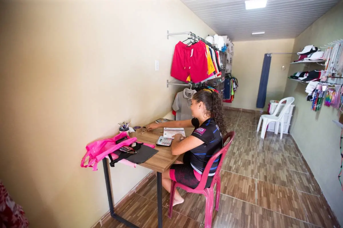 Uma mulher está sentada de costas em uma escrivaninha. Ela tem cabelos cacheados e veste uma camisa de time de futebol