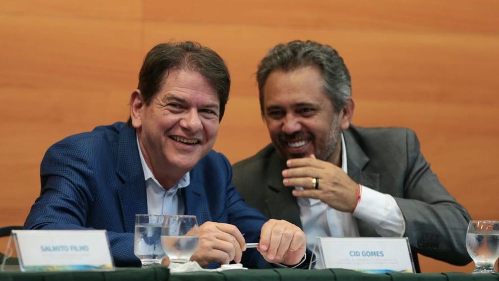 Cid Gomes ao lado de Elmano de Freitas, conversando, sentados à mesa de evento no Pecém