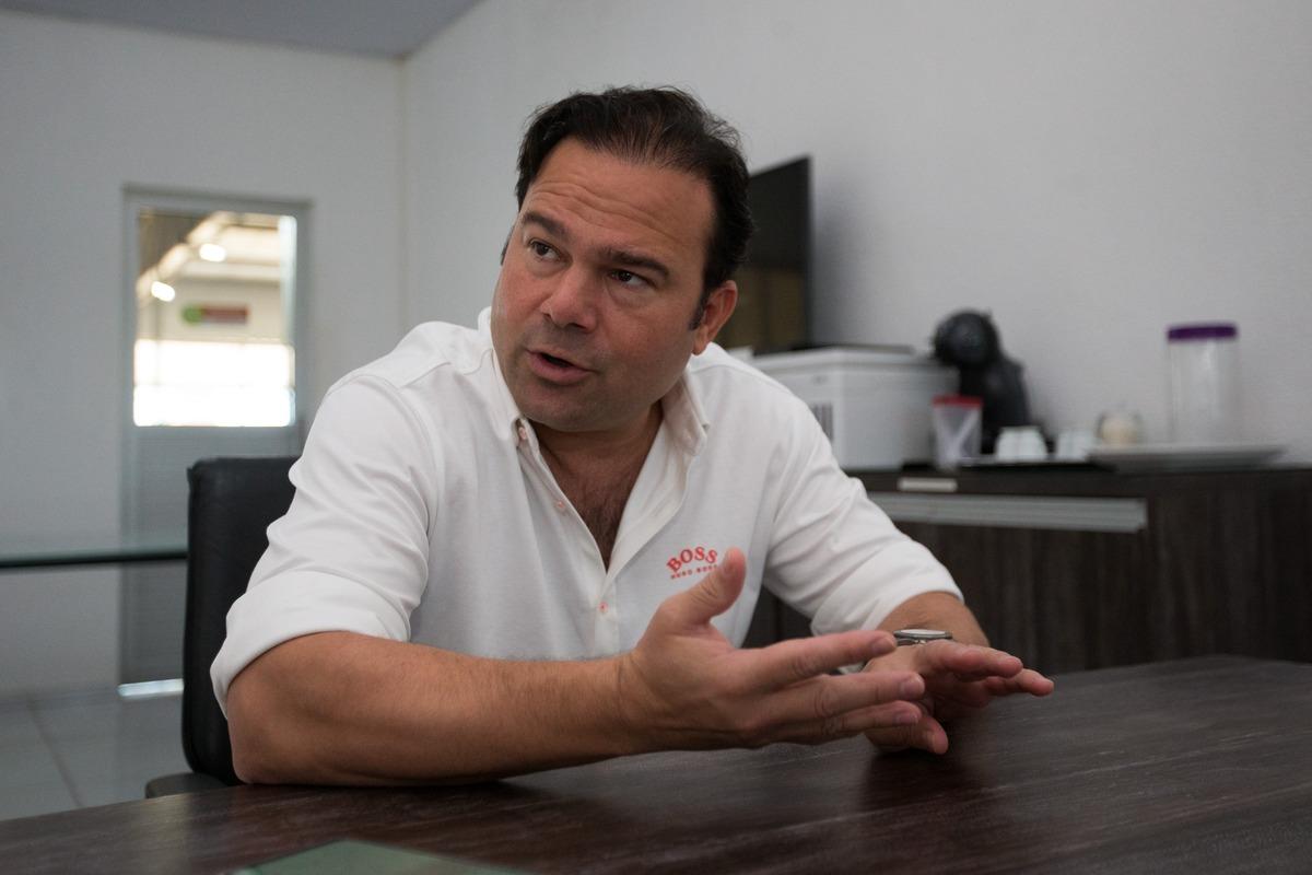 Victor Lima falando em entrevista. Um homem branco, que veste camisa branca, está sentado e gesticulando enquanto conversa com a repórter
