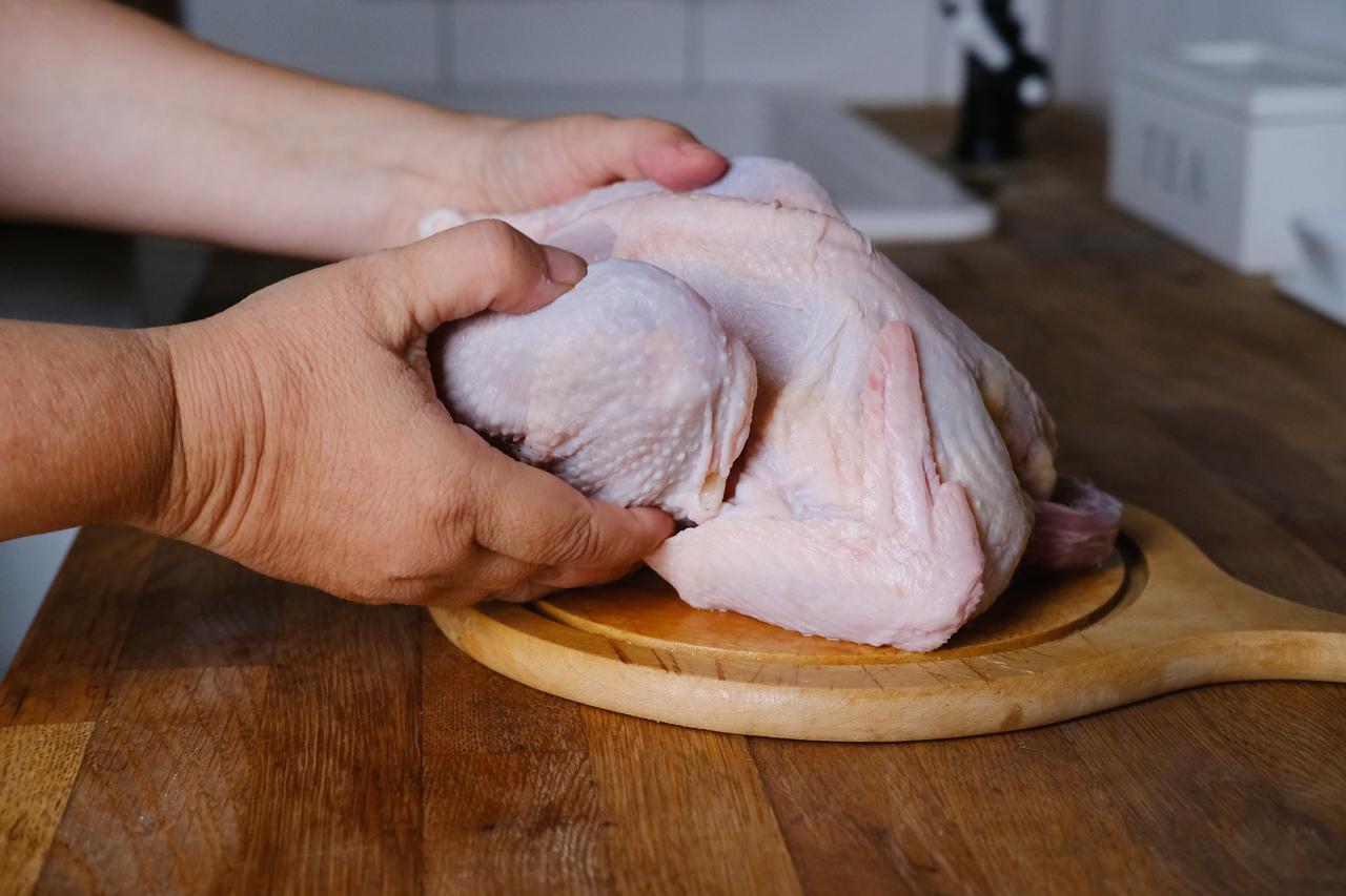 Imagem mostra mãos manipulando frango cru inteiro sobre tábua de corte de madeira, ilustrando se é seguro consumir carnes de aves e ovos após caso de gripe aviária no Brasil