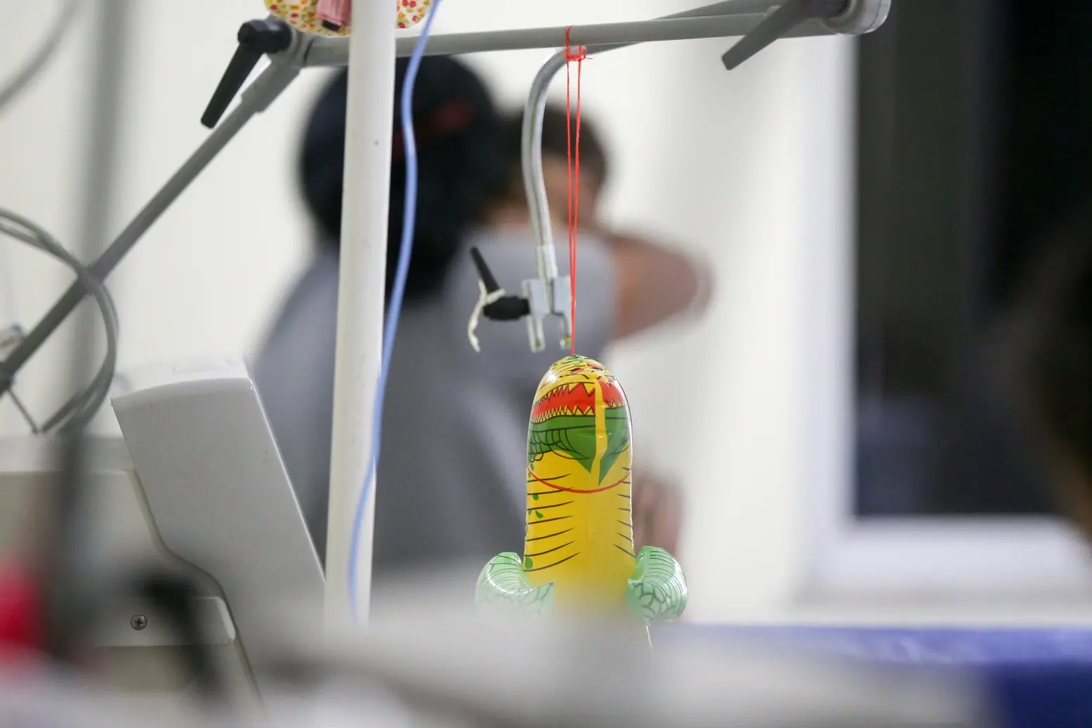 Foto de brinquedo de plástico amarelo em enfermaria infantil; em fundo desfocado, mãe abraça uma criança doente