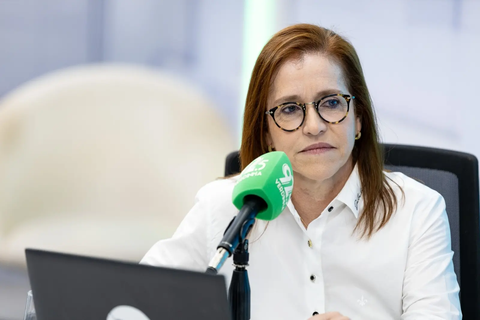 Imagem mostra a secretária da saúde de Fortaleza, uma mulher com cabelos ruivos e óculos de armação redonda escura, fala em um microfone verde com logo da Verdinha FM, em ambiente de estúdio da rádio.