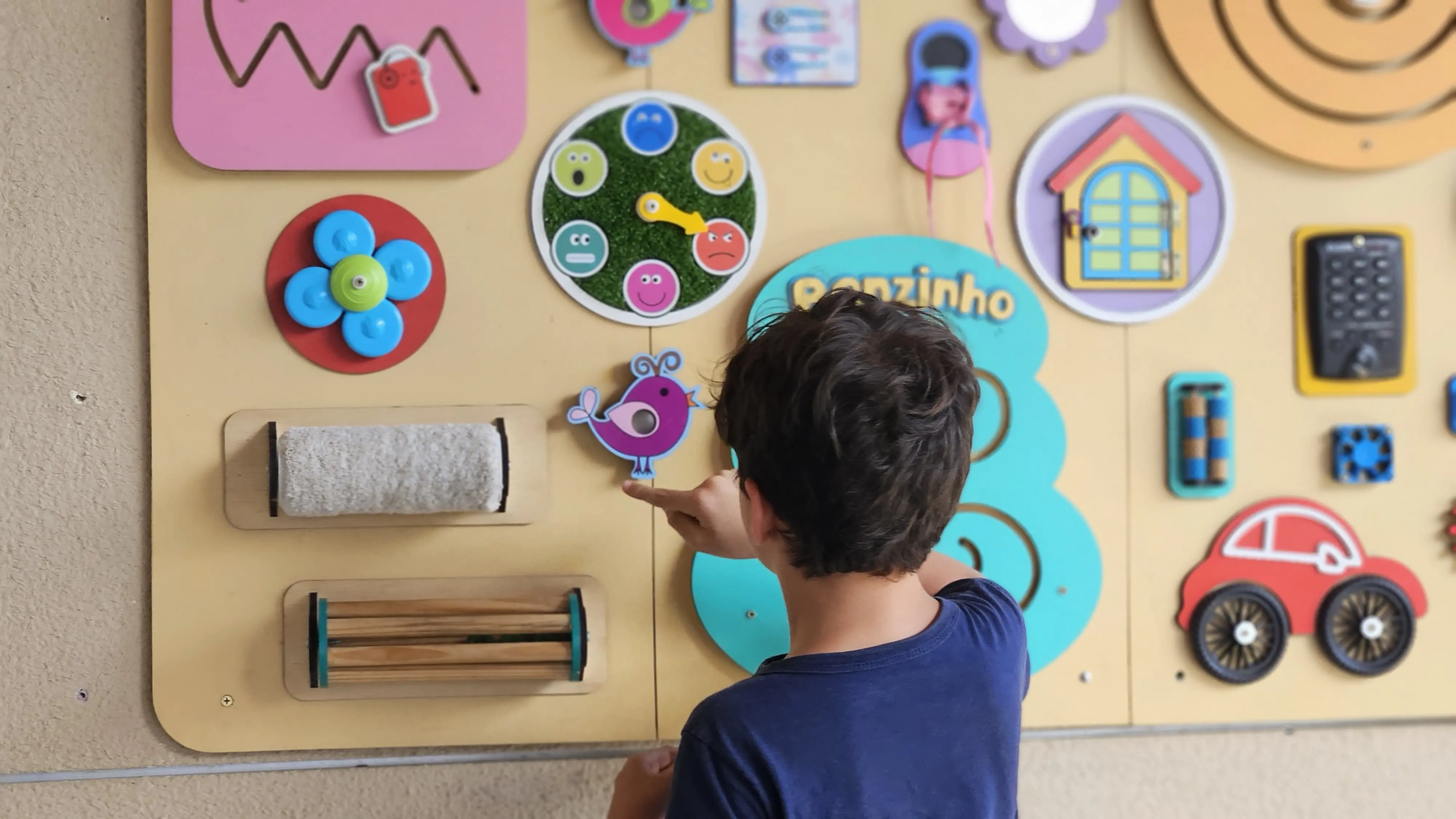 Imagem mostra menino autista cearense de costas, com cabelos pretos lisos e uma camisa azul marinho, apontando para um quadro com diversos brinquedos colados