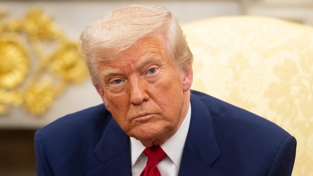 Imagem de Donald Trump com expressão séria, usando terno azul e gravata vermelha, sentado em ambiente formal.