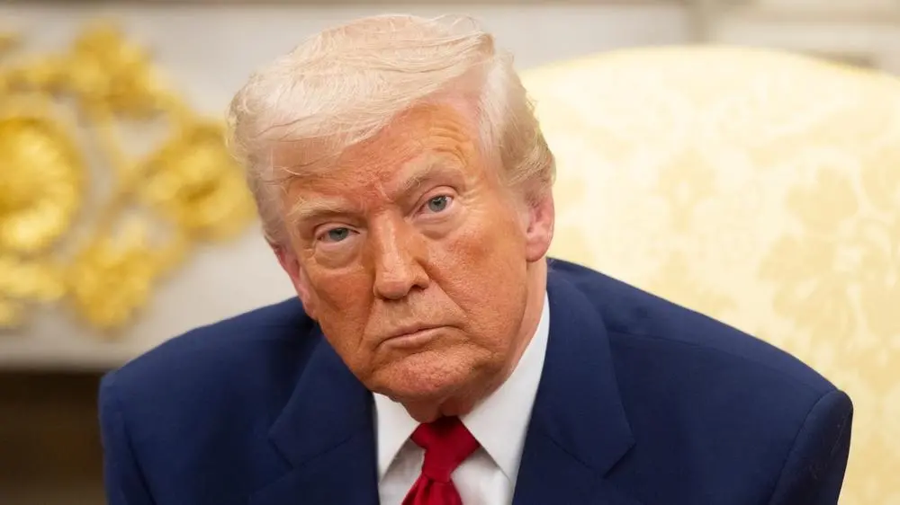 Imagem de Donald Trump com expressão séria, usando terno azul e gravata vermelha, sentado em ambiente formal.