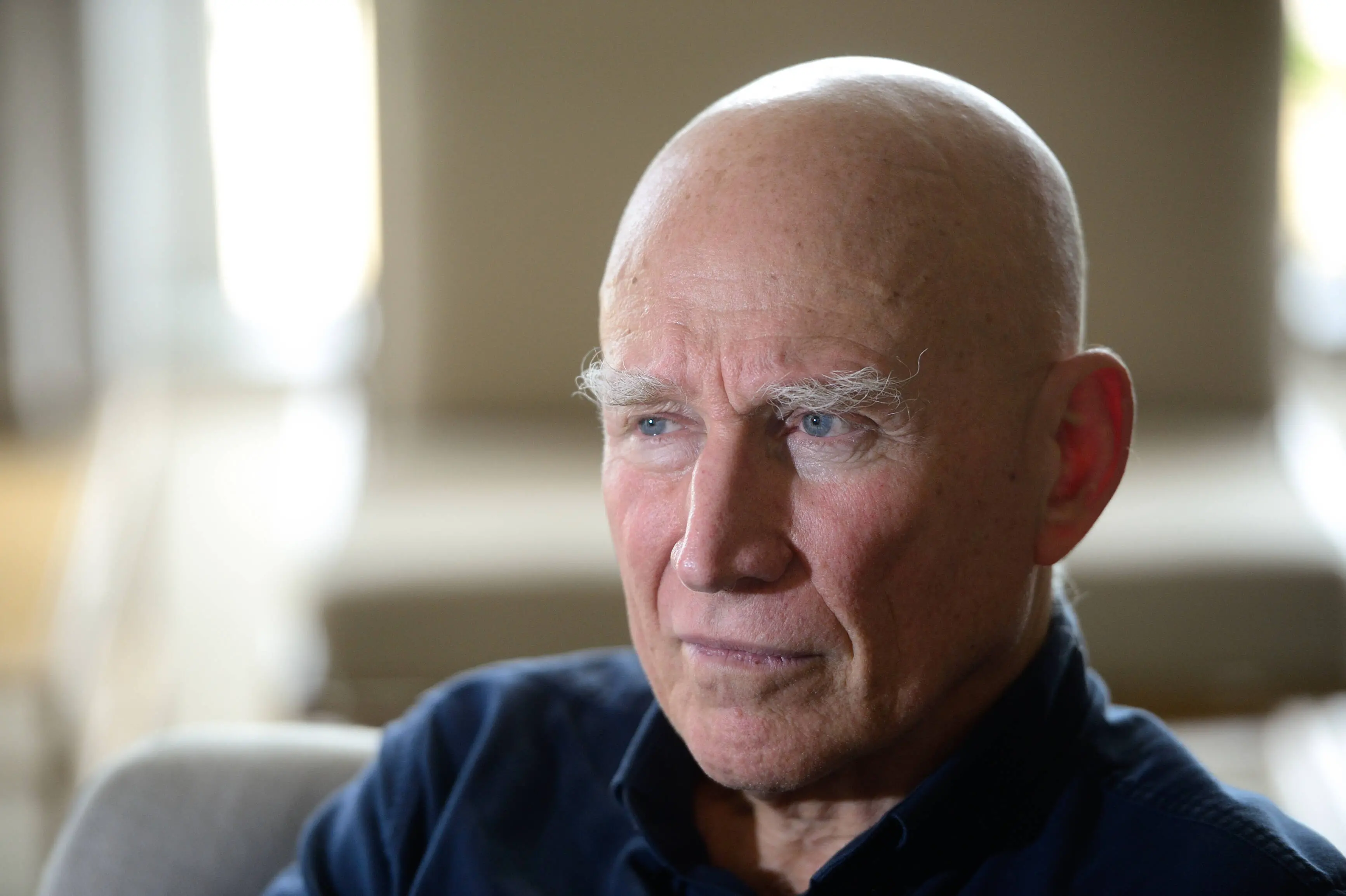 Sebastião Salgado, homem idoso, de pele clara, com sobrancelhas brancas e espessas. Na foto, ele veste uma camisa azul escura e está sentado em um ambiente interno bem iluminado, com fundo desfocado que sugere um ambiente aconchegante. Seu olhar é sério e contemplativo, direcionado para a direita da imagem.