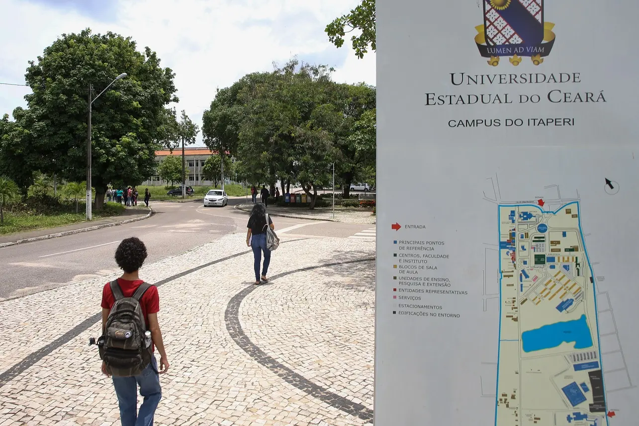 Dois alunos caminham na entrada do campus Itaperi da Uece ao lado de mapa do campus, para reportagem sobre falta de professores na Universidade